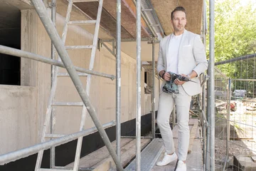 Tomas Sniadowski steht mit einer Bohrmaschine in der Hand auf einem Baugerüst, er trägt einen grauen Anzug.