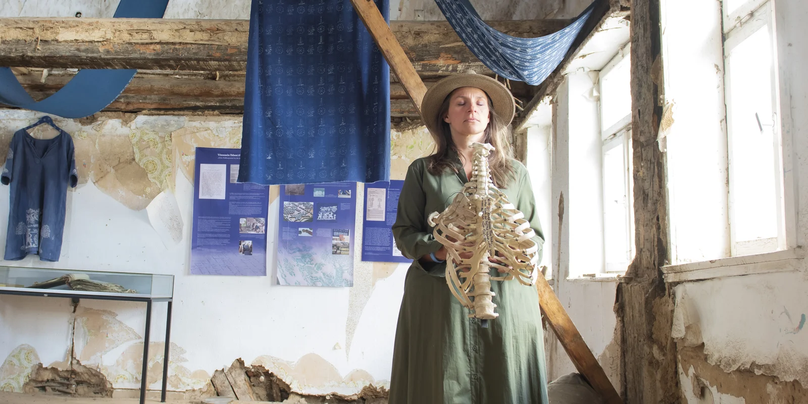 Patricia Magdalene Keil hält den Torso eines Skeletts in den Händen, ihre Augen sind geschlossen. Im Hintergrund ein Fachwerkhaus mit vielen Blaudruck-Stoffen, welche über die Balken gehängt sind.