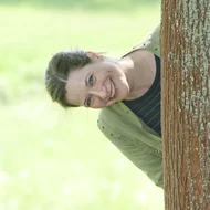 Katrin Fiege schaut seitlich hinter einem großen Baumstamm hervor