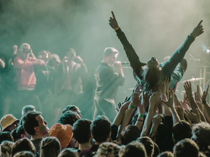 Foto einer Person, die von Konzertbesucher:innen auf Händen getragen wird, im HIntergrund Menschen auf einer Bühne, unten rechts das kulturis-Logo.